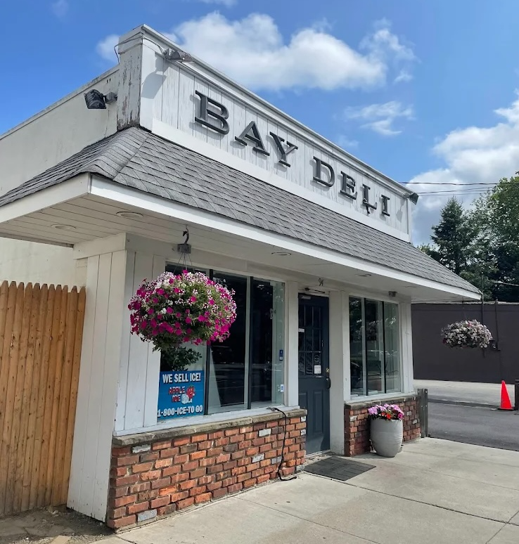 Bay Deli storefront on a sunny day: white facade, gray shingled awning, and entrance between two display windows